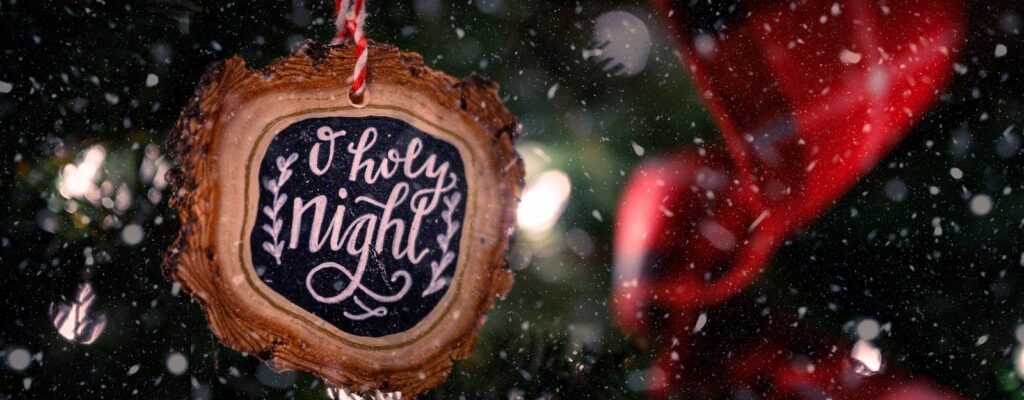Wooden bauble with O Holy Night inscription hanging on green Christmas tree decorated with red ribbon on snowy day