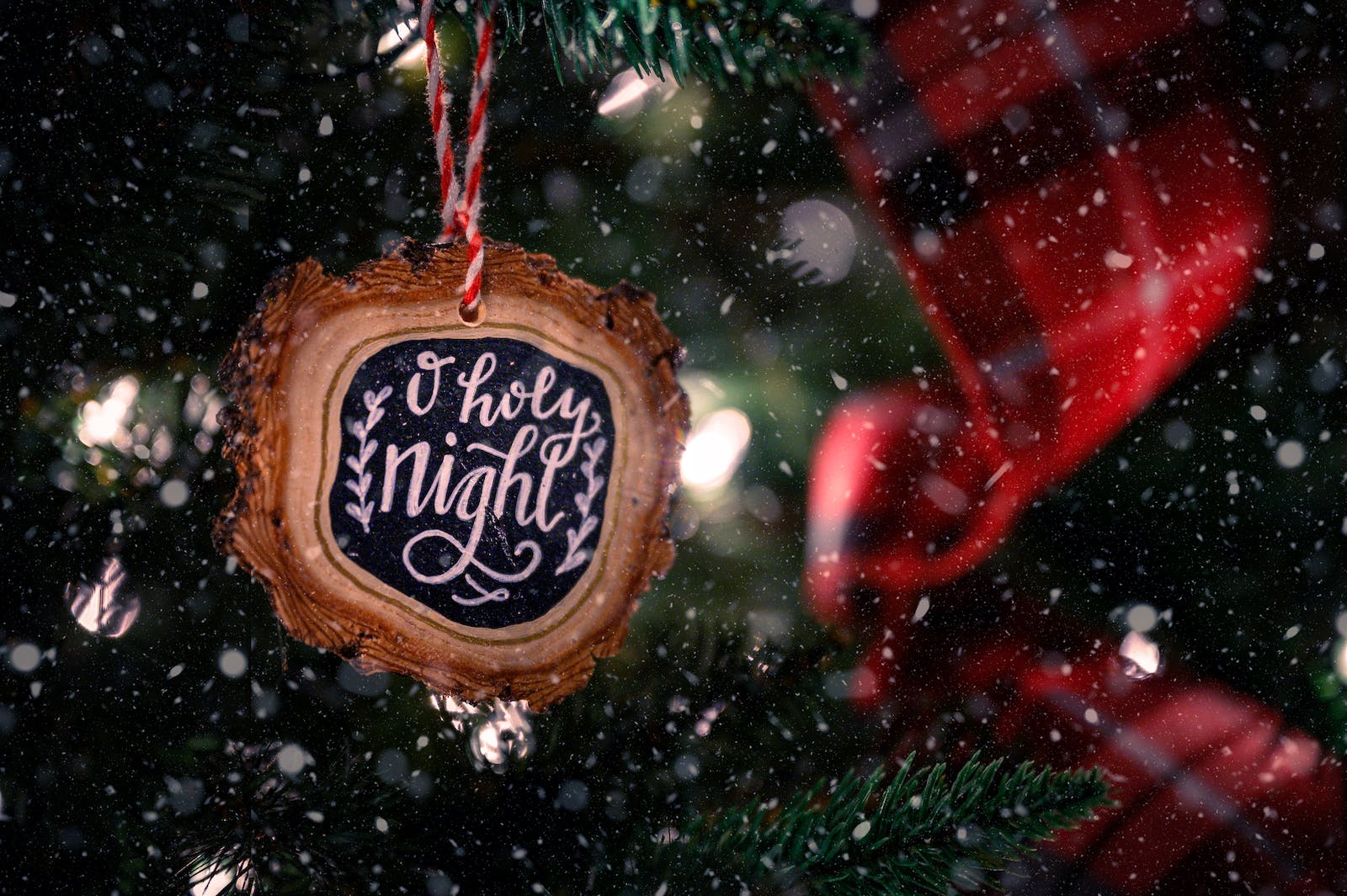 Wooden bauble with O Holy Night inscription hanging on green Christmas tree decorated with red ribbon on snowy day