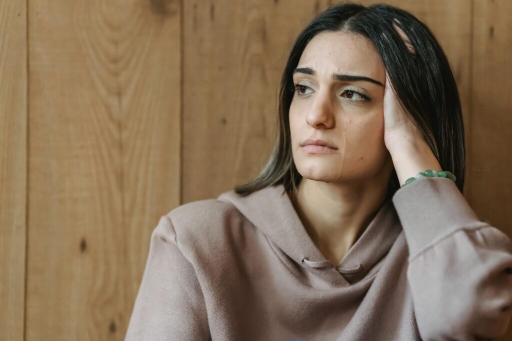 Unhappy young woman with brown hair in casual clothes leaning on hand and looking away while crying in room with wooden wall