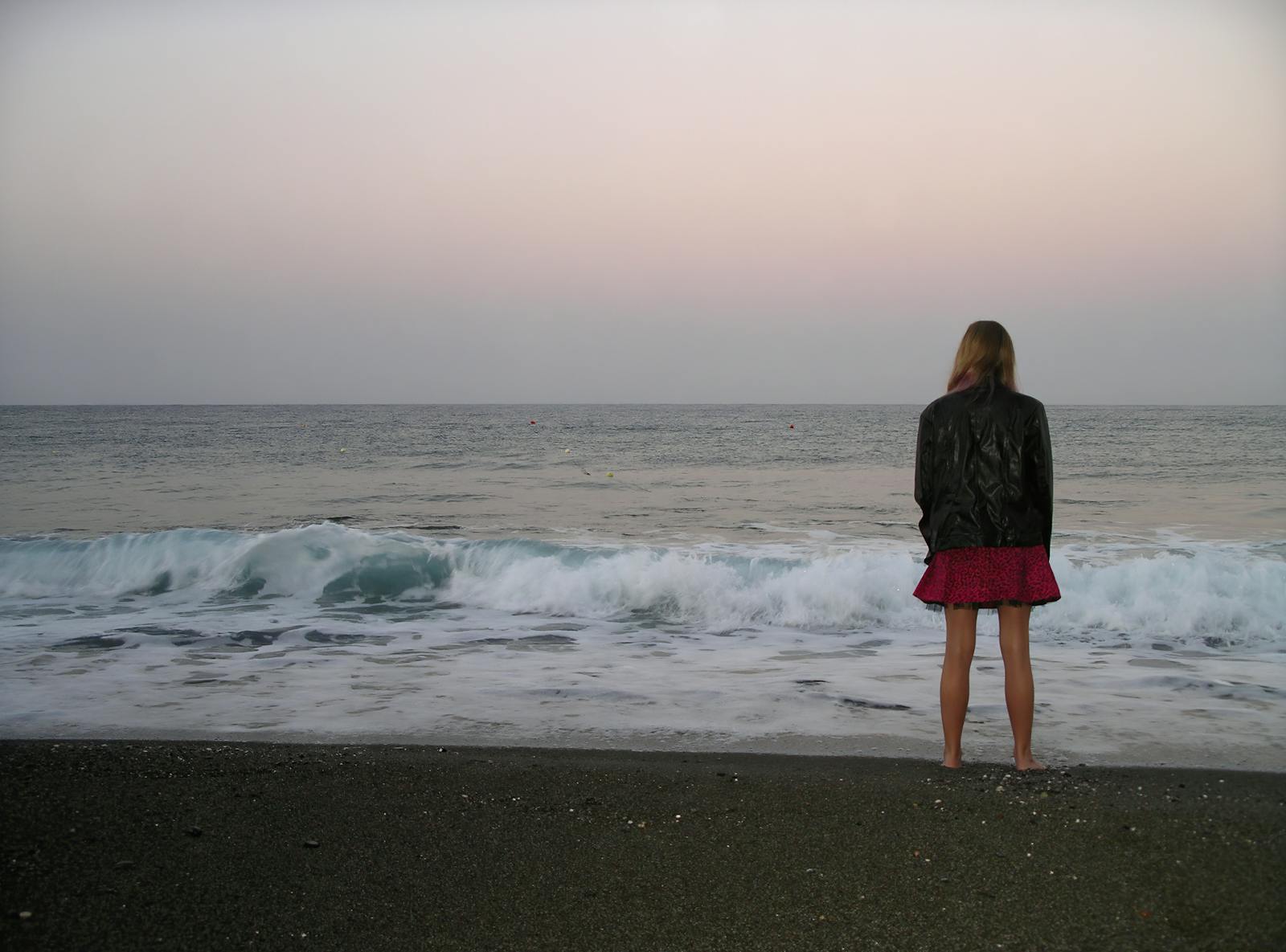 Back View Photo of Barefoot Woman in Black Leather Jacket and Standing by the Beach Looking at the Horizon